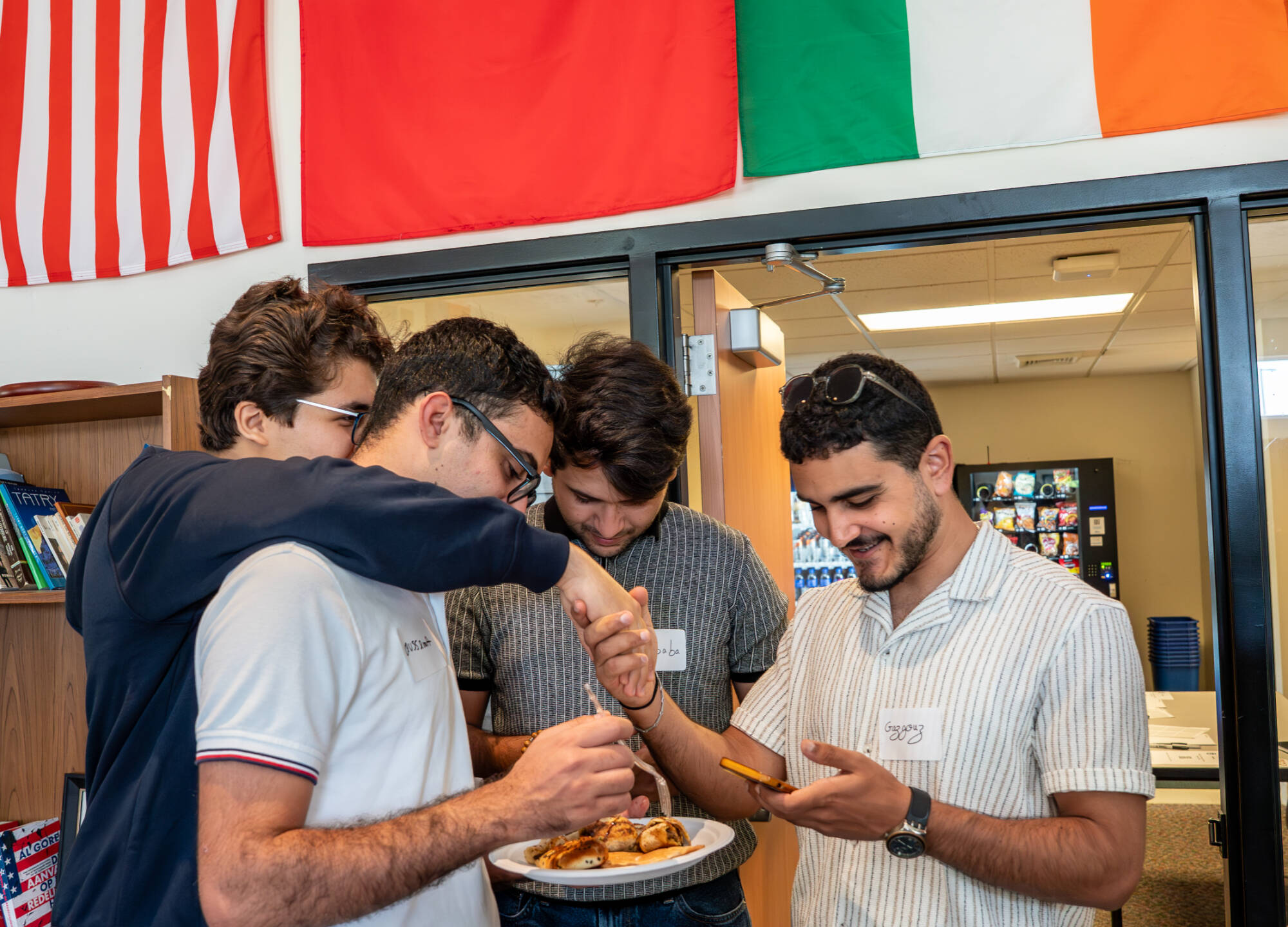 Four students share food and laughter during a College of Computing mixer at GVSU, standing together under international flags and enjoying conversation.